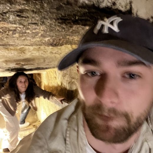 Alec squeezing through a tight tomb tunnel with a friend behind him